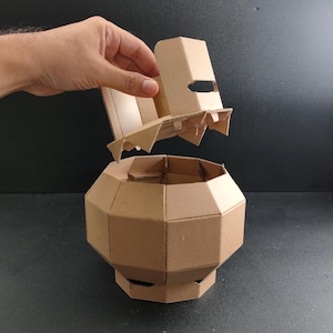 May include: A light brown cardboard container with a faceted, multi-sided geometric body and a matching removable lid. The structure is composed of interlocking panels, featuring a wide, rounded midsection that tapers towards the base.