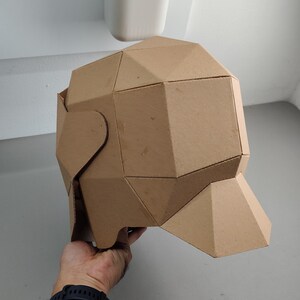 May include: A geometric, brown cardboard sculpture of a head, featuring multiple triangular and rectangular facets. The sculpture is held by a person. The background is a light gray wall and a white object.