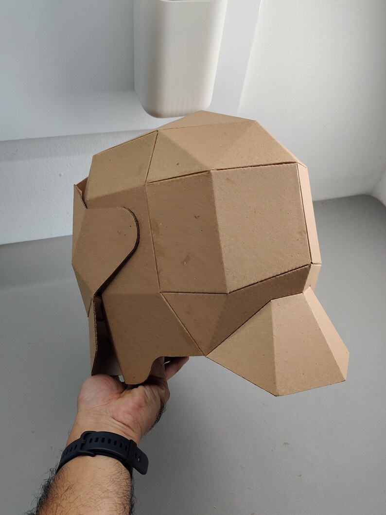 May include: A geometric, brown cardboard sculpture of a head, featuring multiple triangular and rectangular facets. The sculpture is held by a person. The background is a light gray wall and a white object.