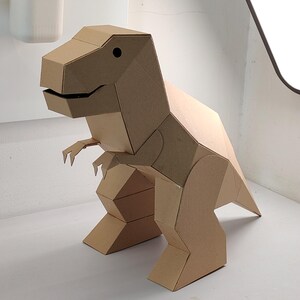 May include: A three-dimensional T-Rex dinosaur figure constructed from light brown cardboard. The model features geometric, angular panels, a large head with a dark circular eye, small arms, and a sturdy tail, showcasing a clean, folded design.