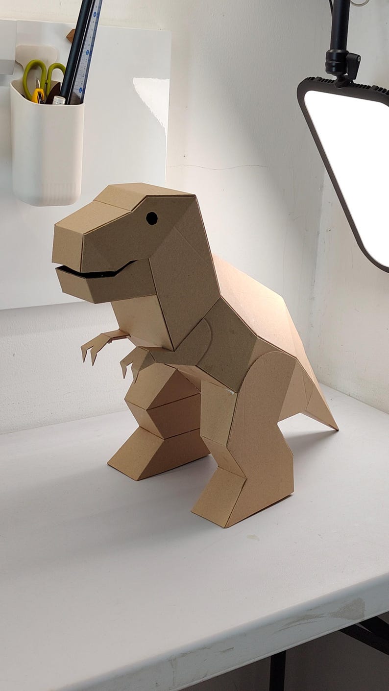 May include: A three-dimensional T-Rex dinosaur figure constructed from light brown cardboard. The model features geometric, angular panels, a large head with a dark circular eye, small arms, and a sturdy tail, showcasing a clean, folded design.