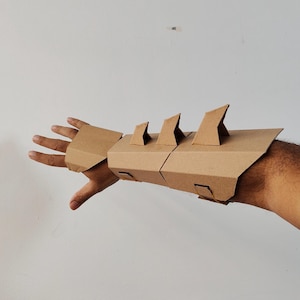 Può includere: Un guanto da braccio di cartone marrone con dettagli angolari e appuntiti. Il guanto è costruito con pezzi di cartone piegati, con una serie di forme triangolari rivolte verso l'alto lungo il bordo superiore. Il design sembra fatto a mano.