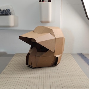Op de afbeelding: Een geometrische helm gemaakt van bruin karton, voorzien van een opvallend vizier en een gefacetteerd, hoekig ontwerp. De structuur is opgebouwd uit meerdere onderling verbonden kartonnen panelen, wat zorgt voor een stijve, polygonale uitstraling.