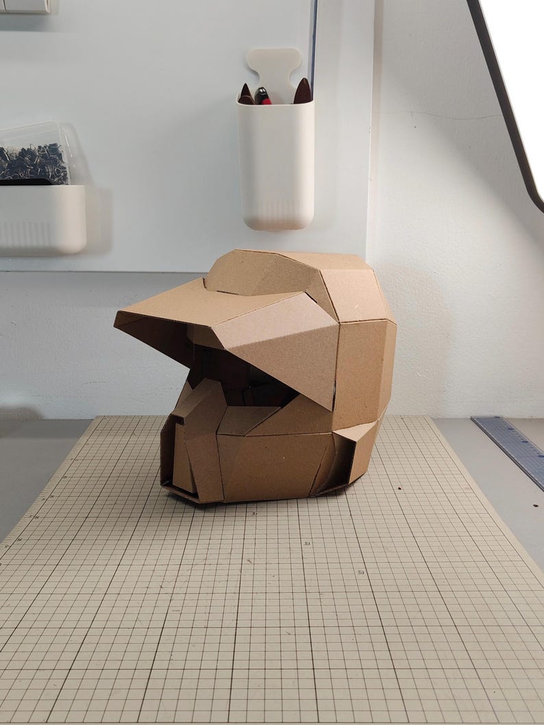 Op de afbeelding: Een geometrische helm gemaakt van bruin karton, voorzien van een opvallend vizier en een gefacetteerd, hoekig ontwerp. De structuur is opgebouwd uit meerdere onderling verbonden kartonnen panelen, wat zorgt voor een stijve, polygonale uitstraling.