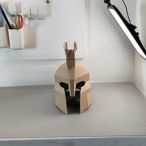 May include: A tan cardboard helmet featuring a geometric, angular design with a central crest on top. The helmet has a wide, open face opening with a nose guard and eye cutouts, constructed from multiple flat, folded panels of brown paper material.