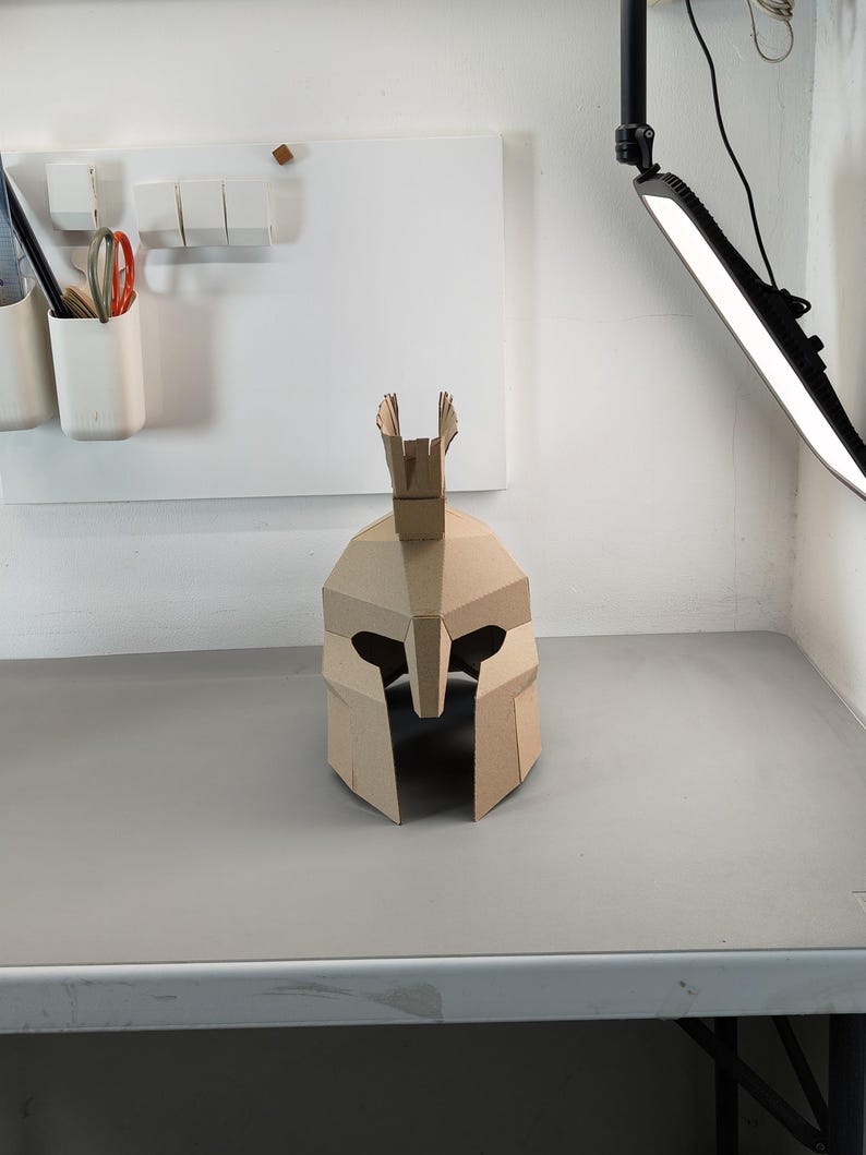 May include: A tan cardboard helmet featuring a geometric, angular design with a central crest on top. The helmet has a wide, open face opening with a nose guard and eye cutouts, constructed from multiple flat, folded panels of brown paper material.