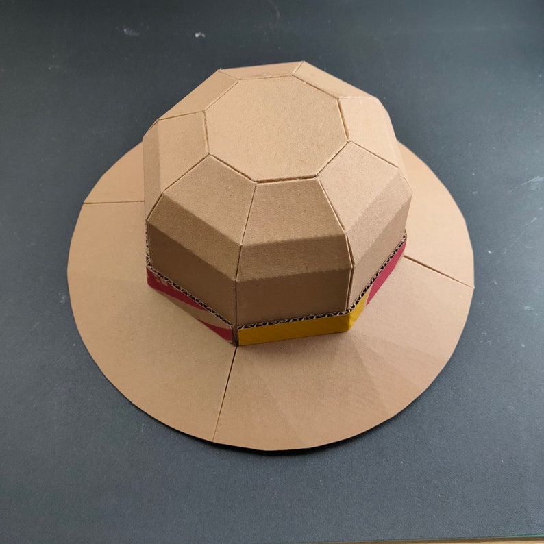 Op de afbeelding: Een kartonnen hoed met een bruin, rood en geel gestreept band. De hoed is gemaakt van meerdere kartonnen stukken die aan elkaar zijn geplakt om een geometrische vorm te cre&euml;ren.