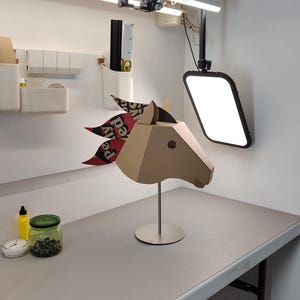Puede incluir: Una escultura geom&eacute;trica de cabeza de caballo hecha de cart&oacute;n color canela, montada sobre un soporte de metal plateado. El caballo presenta una crin estilizada hecha de papel estampado en capas con elementos de texto en rojo, negro y blanco, incluyendo "pely".