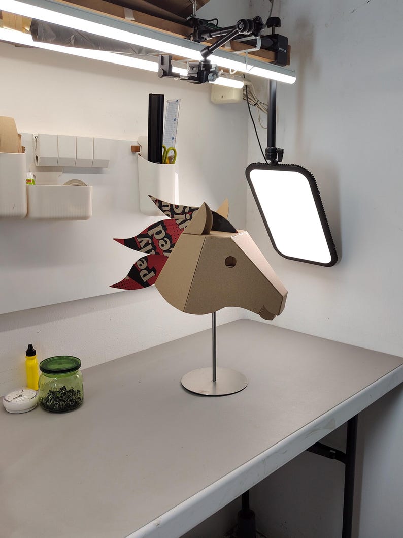 Puede incluir: Una escultura geom&eacute;trica de cabeza de caballo hecha de cart&oacute;n color canela, montada sobre un soporte de metal plateado. El caballo presenta una crin estilizada hecha de papel estampado en capas con elementos de texto en rojo, negro y blanco, incluyendo "pely".