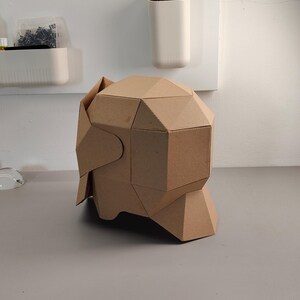 May include: A geometric, cardboard sculpture of a head, featuring angular planes and a natural brown color. The sculpture is constructed from folded cardboard pieces, creating a unique, modern design. It sits on a gray surface.