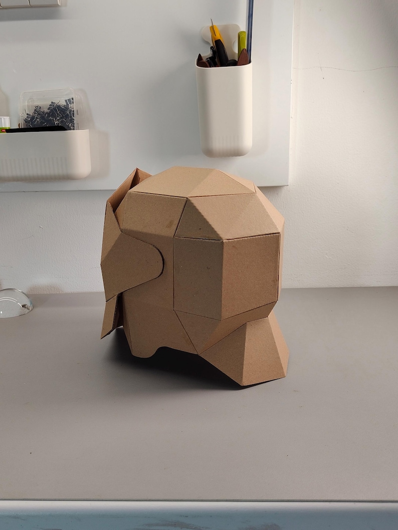 May include: A geometric, cardboard sculpture of a head, featuring angular planes and a natural brown color. The sculpture is constructed from folded cardboard pieces, creating a unique, modern design. It sits on a gray surface.