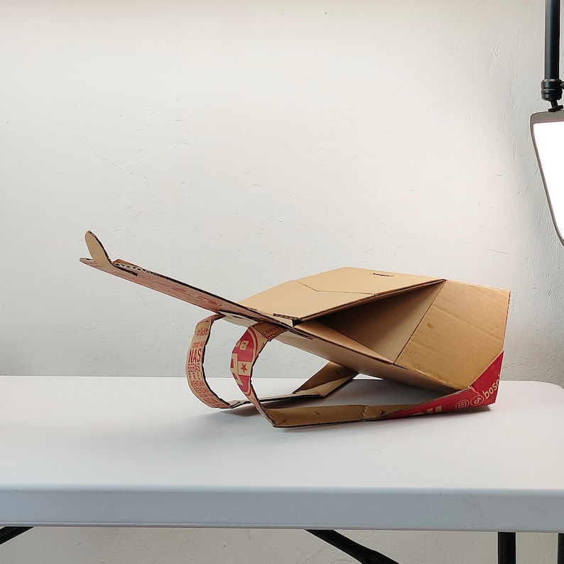 May include: A cardboard pizza box has been folded into a unique shape, resembling a futuristic vehicle. The box is brown with red and white text and a star logo.