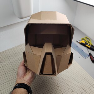 Op de afbeelding: Een geometrische helm gemaakt van bruin golfkarton. De helm heeft een brede vizieropening, een opvallend hoekig neusgedeelte en zijpanelen. Het ontwerp is samengesteld uit meerdere platte, gevouwen kartonnen segmenten.