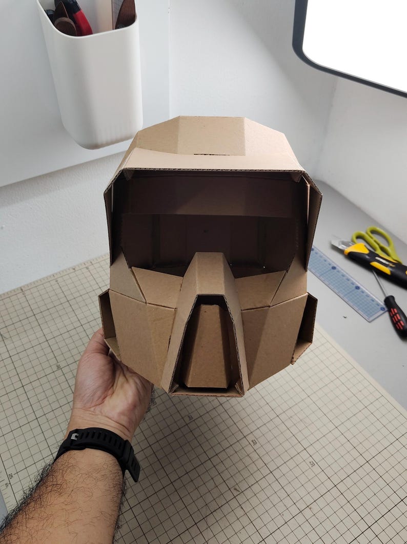 Op de afbeelding: Een geometrische helm gemaakt van bruin golfkarton. De helm heeft een brede vizieropening, een opvallend hoekig neusgedeelte en zijpanelen. Het ontwerp is samengesteld uit meerdere platte, gevouwen kartonnen segmenten.