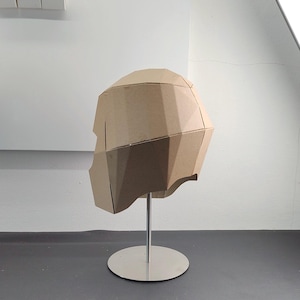 May include: A geometric, low-poly style helmet constructed from light brown cardboard panels. The helmet features a faceted, angular design and is mounted on a silver-coloured metal display stand with a circular base.