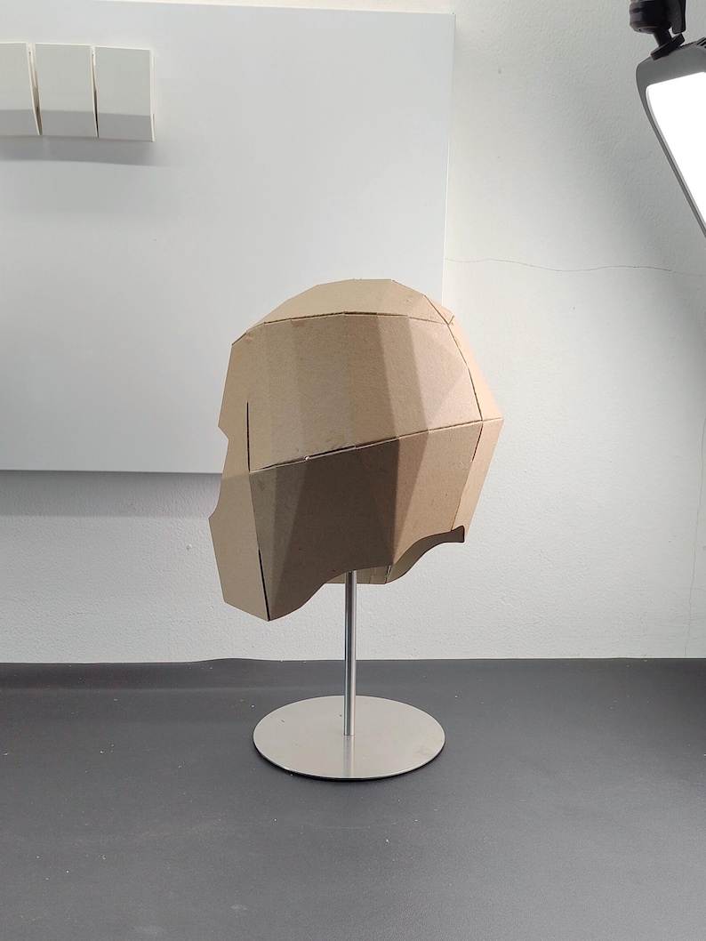May include: A geometric, low-poly style helmet constructed from light brown cardboard panels. The helmet features a faceted, angular design and is mounted on a silver-coloured metal display stand with a circular base.