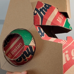 May include: A geometric structure crafted from brown cardboard, featuring a circular cutout that reveals a red, green, and white printed surface with the text Mi Instan. The design incorporates angular panels and exposed corrugated edges.