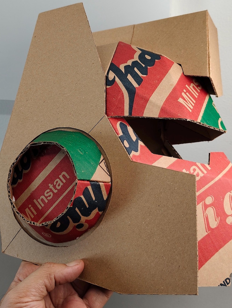 May include: A geometric structure crafted from brown cardboard, featuring a circular cutout that reveals a red, green, and white printed surface with the text Mi Instan. The design incorporates angular panels and exposed corrugated edges.