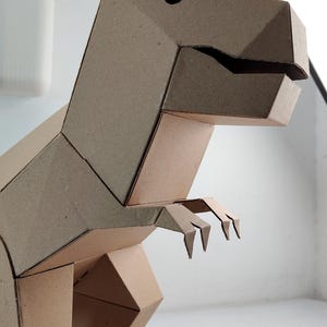 May include: A three-dimensional T-Rex dinosaur figure constructed from brown cardboard. The model features a geometric design with angular panels, a distinct head with a cutout eye, a wide jaw, and two small arms extending from the torso.