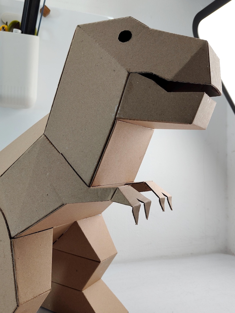 May include: A three-dimensional T-Rex dinosaur figure constructed from brown cardboard. The model features a geometric design with angular panels, a distinct head with a cutout eye, a wide jaw, and two small arms extending from the torso.