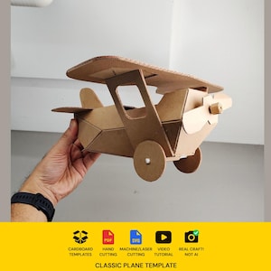 May include: A brown cardboard biplane model featuring a propeller, two wings, and circular wheels. The base of the image includes text: CARDBOARD TEMPLATES, PDF, SVG, MACHINE/LASER CUTTING, VIDEO TUTORIAL, REAL CRAFT! NOT AI, CLASSIC PLANE TEMPLATE.