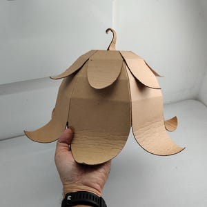 May include: A bell-shaped structure constructed from brown corrugated cardboard, featuring multiple overlapping petal-like segments arranged in two tiers and a small curved hook at the top center, designed to resemble a flower or bell.
