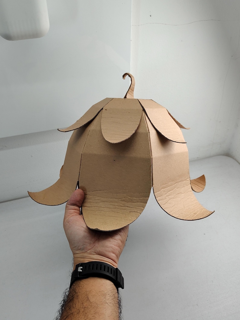 May include: A bell-shaped structure constructed from brown corrugated cardboard, featuring multiple overlapping petal-like segments arranged in two tiers and a small curved hook at the top center, designed to resemble a flower or bell.