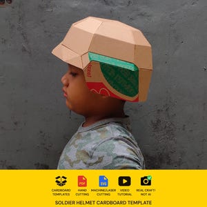 Pode incluir: Um capacete de soldado de papelão bege com um design geométrico e facetado. A lateral do capacete apresenta seções de papelão impressas em verde e vermelho. O capacete é construído a partir de painéis dobrados, criando uma aparência estruturada.