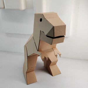 May include: A three-dimensional dinosaur figure constructed from light brown corrugated cardboard. The model features a geometric design with a large head, a wide jaw, two small arms, and two sturdy legs, all assembled with visible folded edges.