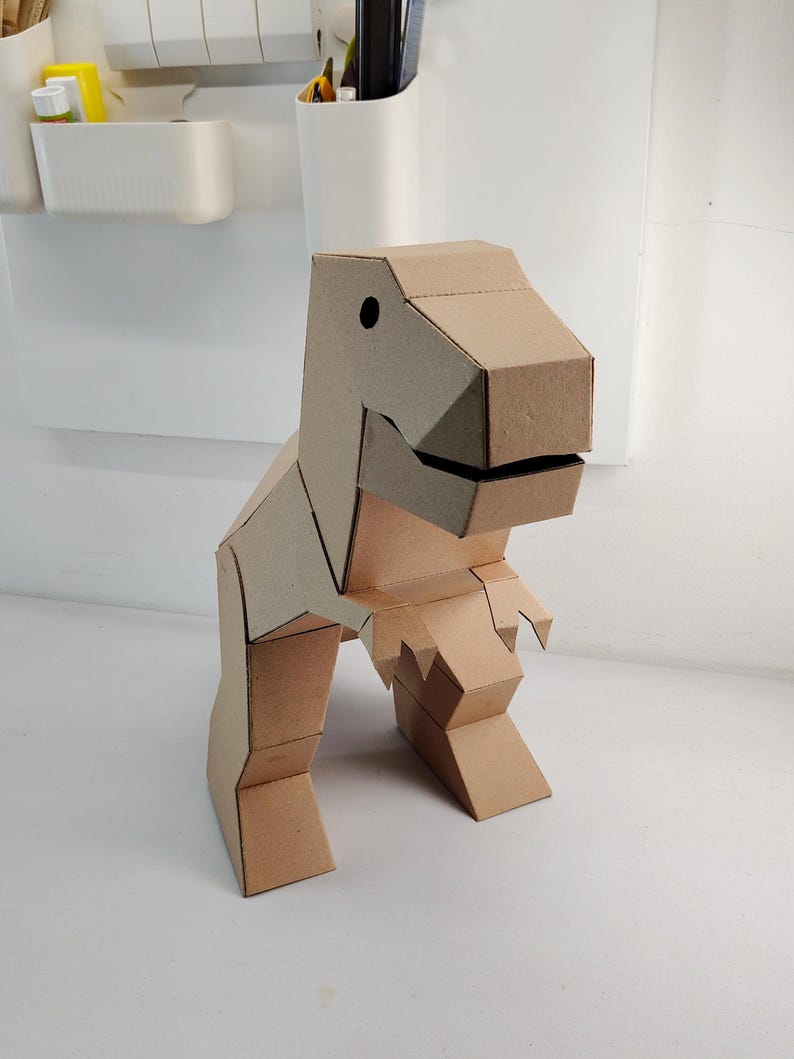 May include: A three-dimensional dinosaur figure constructed from light brown corrugated cardboard. The model features a geometric design with a large head, a wide jaw, two small arms, and two sturdy legs, all assembled with visible folded edges.