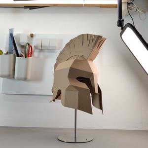 May include: A tan cardboard Spartan-style helmet featuring a high crest and angular geometric construction. The helmet is mounted on a silver-colored metal stand with a circular base, showcasing a detailed layered design with visible seams.