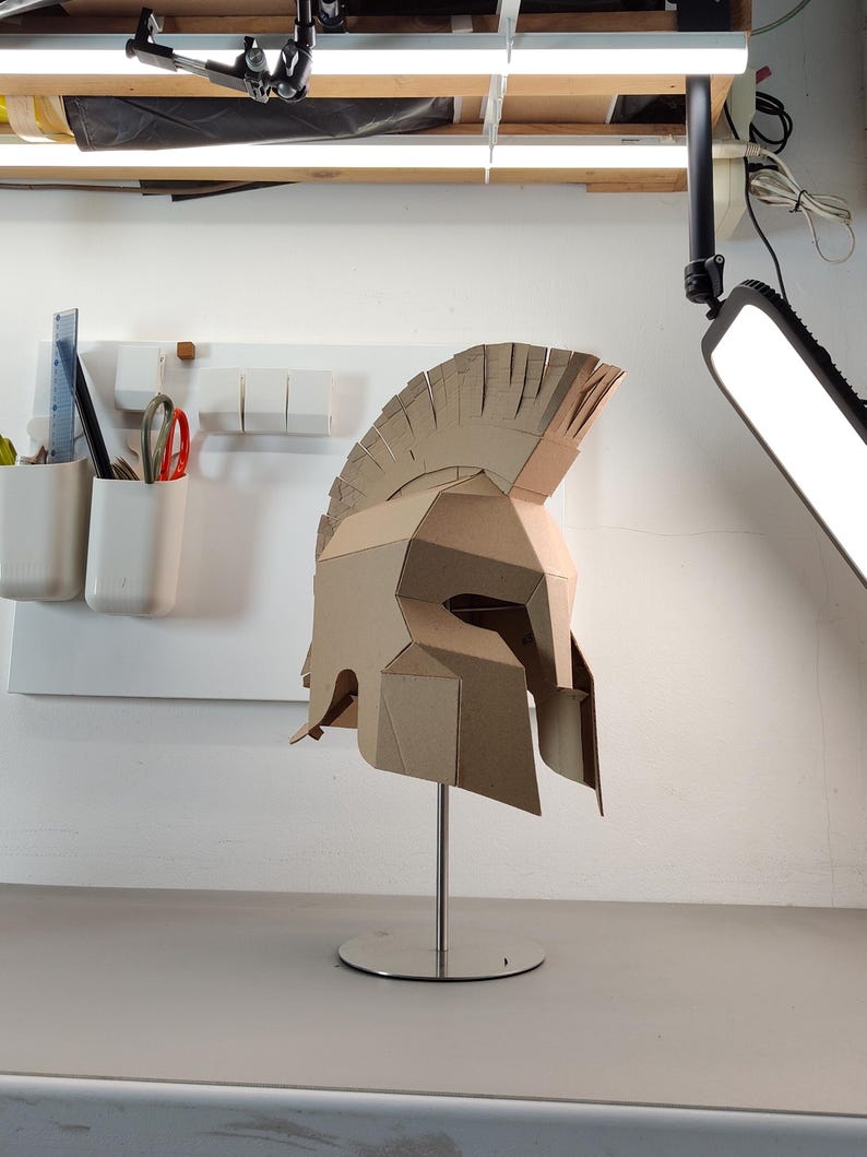 May include: A tan cardboard Spartan-style helmet featuring a high crest and angular geometric construction. The helmet is mounted on a silver-colored metal stand with a circular base, showcasing a detailed layered design with visible seams.