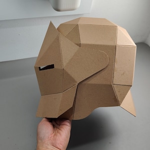 May include: A geometric cardboard rhino mask, featuring angular planes and a cutout for the eye. The mask is held by a person's hand, showcasing its three-dimensional design and craft paper construction.