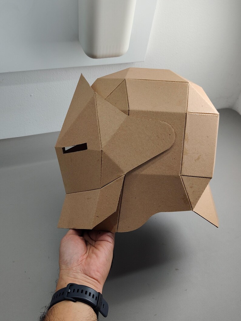 May include: A geometric cardboard rhino mask, featuring angular planes and a cutout for the eye. The mask is held by a person's hand, showcasing its three-dimensional design and craft paper construction.