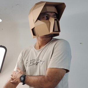 Op de afbeelding: Een geometrische helm gemaakt van bruin karton met een vizier en een gestructureerde voorplaat met hoekige facetten. De helm heeft een rigide ontwerp met meerdere panelen die het hoofd en gezicht bedekken, met een duidelijke low-poly esthetiek.