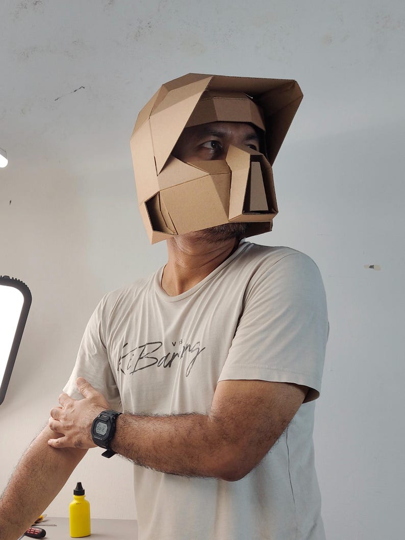 Op de afbeelding: Een geometrische helm gemaakt van bruin karton met een vizier en een gestructureerde voorplaat met hoekige facetten. De helm heeft een rigide ontwerp met meerdere panelen die het hoofd en gezicht bedekken, met een duidelijke low-poly esthetiek.