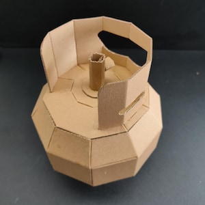 May include: A geometric model of a gas cylinder constructed from light brown cardboard. The structure features a faceted, multi-sided body, a central vertical tube, and a protective handle assembly at the top, all crafted with visible seams.