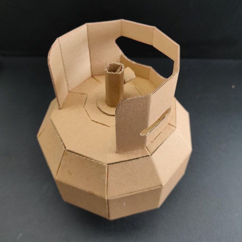 May include: A geometric model of a gas cylinder constructed from light brown cardboard. The structure features a faceted, multi-sided body, a central vertical tube, and a protective handle assembly at the top, all crafted with visible seams.