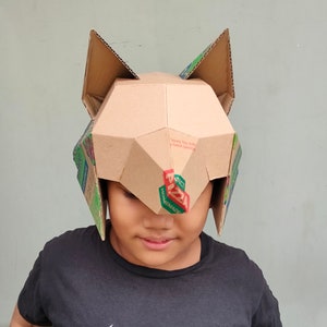 May include: A handmade cardboard fox mask with geometric shapes and pointed ears. The mask is constructed from recycled cardboard boxes, with visible text and graphics from the original packaging. The mask covers the top of the head and forehead.