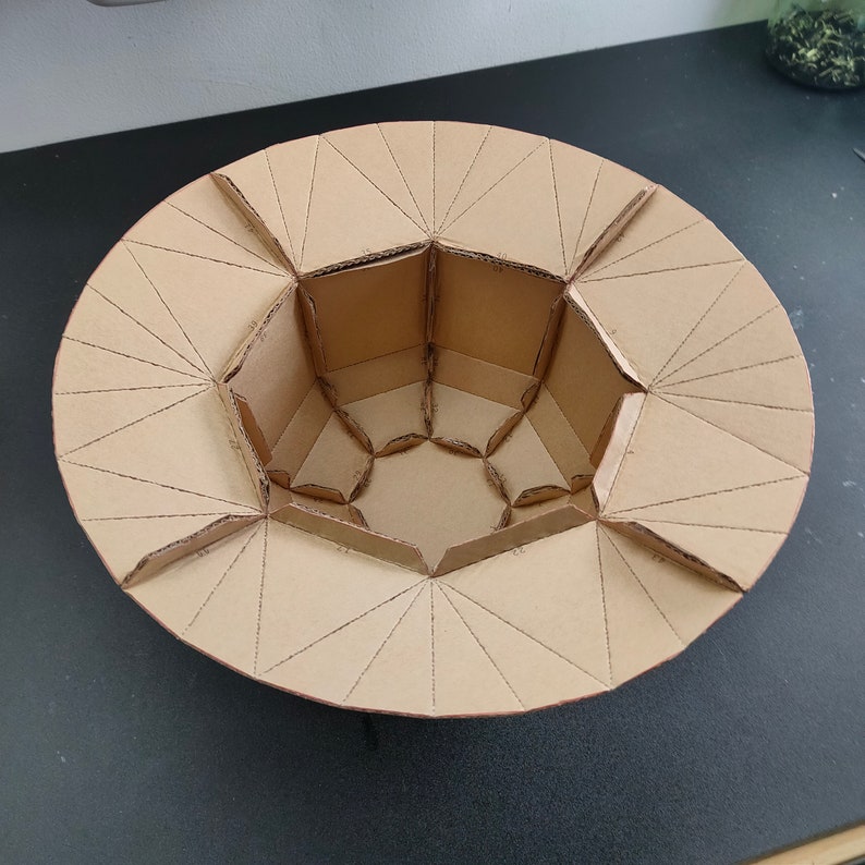 Op de afbeelding: Een kartonnen model van een geometrische vorm met een zeshoekige basis en een ronde bovenkant. Het model is gemaakt van bruin karton en is gemarkeerd en gevouwen om de vorm te cre&euml;ren.