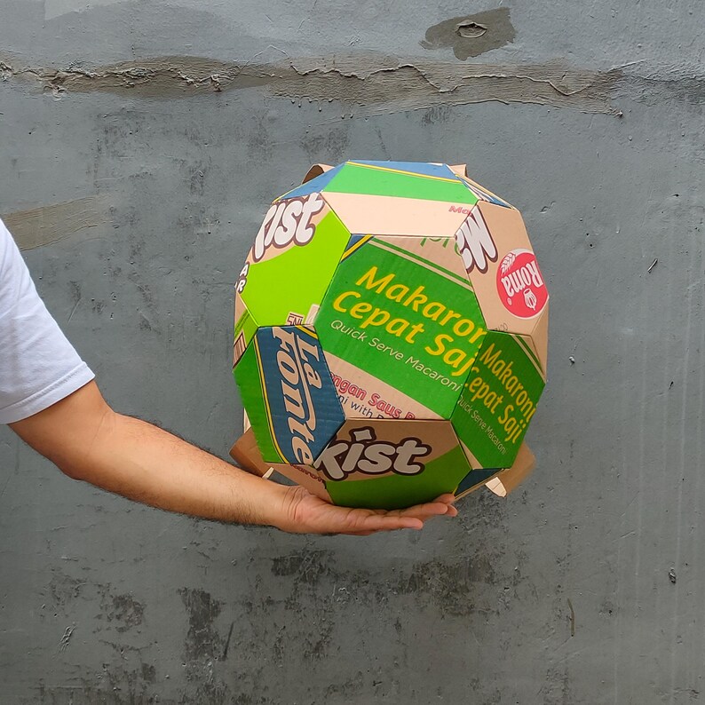 K&ouml;nnte beinhalten: Ein Ball aus Pappe, der aus verschiedenen Lebensmittelverpackungen hergestellt wurde. Der Ball ist mit gr&uuml;nen, roten, blauen und gelben Farben verziert. Der Text auf dem Ball lautet "Makaroni Cepat Saji" und "Quick Serve Macaroni".