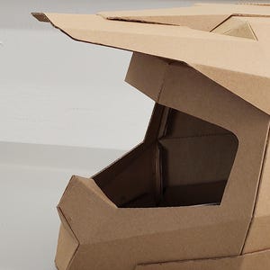 May include: A tan cardboard helmet featuring a geometric, angular design with a prominent visor and a large open face area. The structure is constructed from multiple folded panels of brown corrugated cardboard, showcasing sharp edges and clean lines.