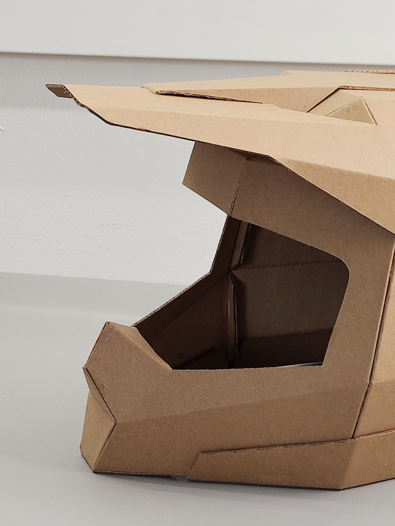 May include: A tan cardboard helmet featuring a geometric, angular design with a prominent visor and a large open face area. The structure is constructed from multiple folded panels of brown corrugated cardboard, showcasing sharp edges and clean lines.