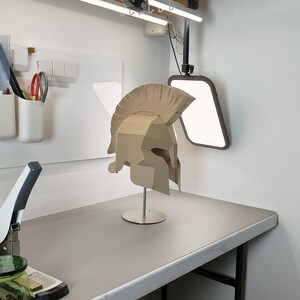 May include: A tan cardboard Spartan-style helmet featuring a prominent crest and angular face guard, mounted on a thin silver metal stand with a circular base. The helmet is constructed from multiple joined pieces of corrugated paper material.