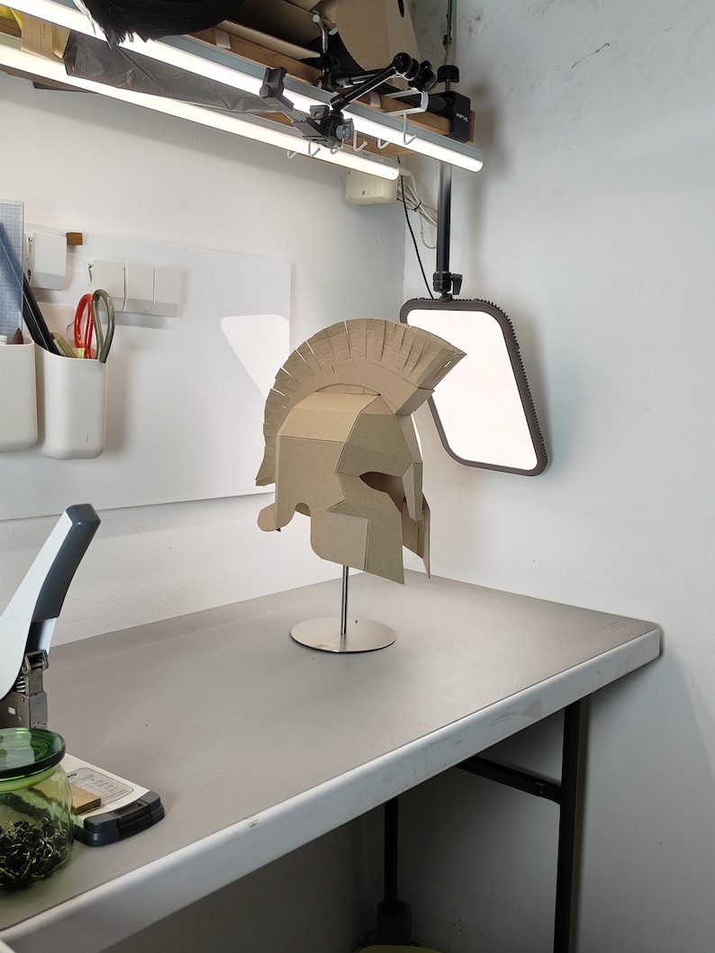 May include: A tan cardboard Spartan-style helmet featuring a prominent crest and angular face guard, mounted on a thin silver metal stand with a circular base. The helmet is constructed from multiple joined pieces of corrugated paper material.
