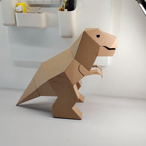 May include: A three-dimensional geometric dinosaur figure constructed from brown cardboard. The model features a large head with a visible eye cutout, a long pointed tail, and two small front limbs, all assembled with clean, angular edges and flat surfaces.