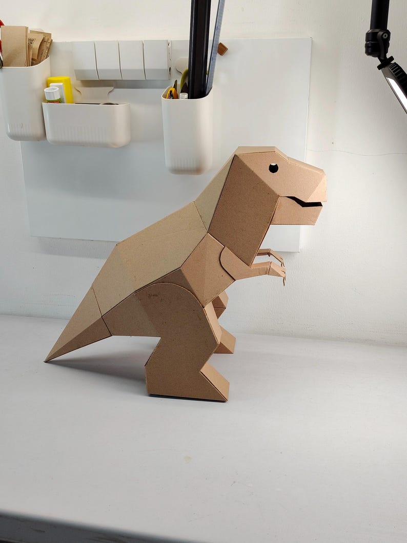 May include: A three-dimensional geometric dinosaur figure constructed from brown cardboard. The model features a large head with a visible eye cutout, a long pointed tail, and two small front limbs, all assembled with clean, angular edges and flat surfaces.