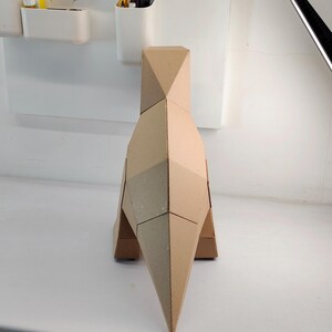 May include: A geometric sculpture of a dinosaur constructed from tan cardboard. The figure features a faceted, angular design with a pointed tail, a tall body, and a narrow neck, showcasing clean, sharp edges and a minimalist, monochromatic appearance.