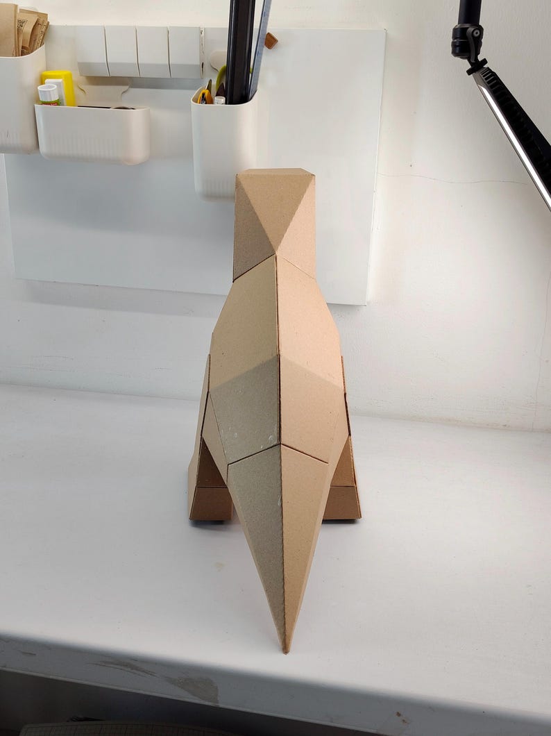 May include: A geometric sculpture of a dinosaur constructed from tan cardboard. The figure features a faceted, angular design with a pointed tail, a tall body, and a narrow neck, showcasing clean, sharp edges and a minimalist, monochromatic appearance.