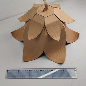 May include: A brown cardboard sculpture shaped like a flower with layered petals and a central stem, positioned behind a clear plastic ruler that measures approximately 11.5 inches in length, showing the object's scale on a white surface.